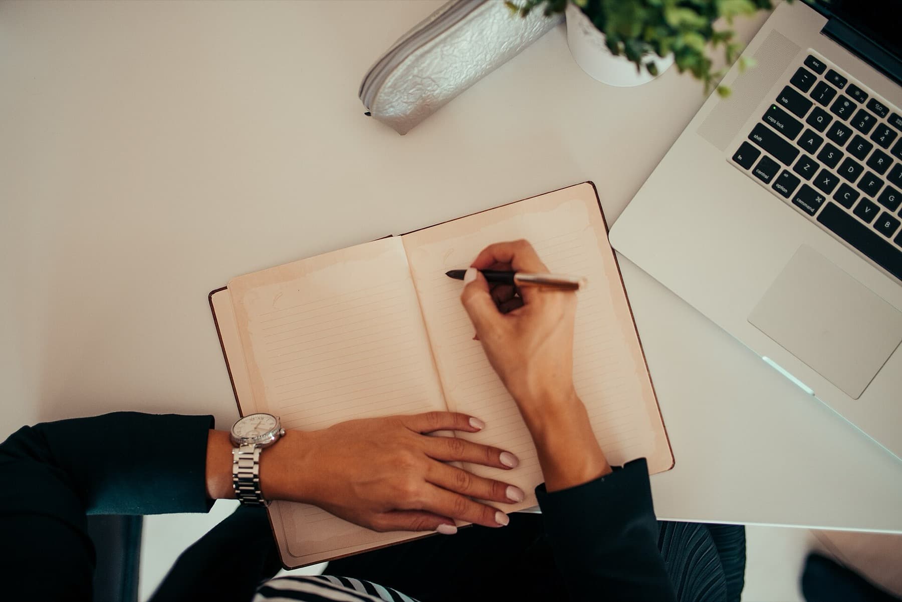 A person writing in a notebook beside a laptop at a working desk.